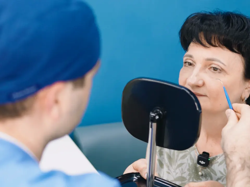Хирург RB CLINIC размечает веко пациентки перед блефаропластикой, пациентка смотрит в зеркало